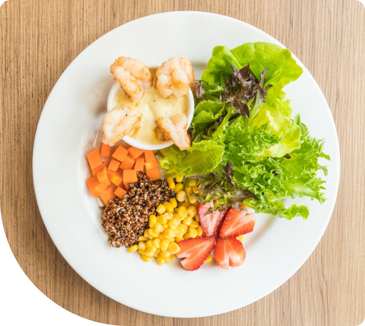 En färgglad sallad med grillade räkor i en vit sås, serverad tillsammans med quinoa, morötter, majs, jordgubbar och olika sorters gröna sallader, allt på en vit tallrik.